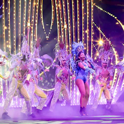 Performers in vibrant costumes and headdresses dancing on a stage with bright lights and a smoky atmosphere.