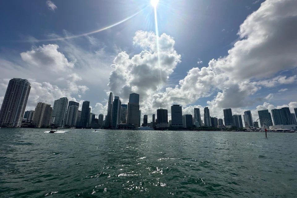 City skyline with tall buildings under a bright sun and partly cloudy sky, viewed from across a body of water.