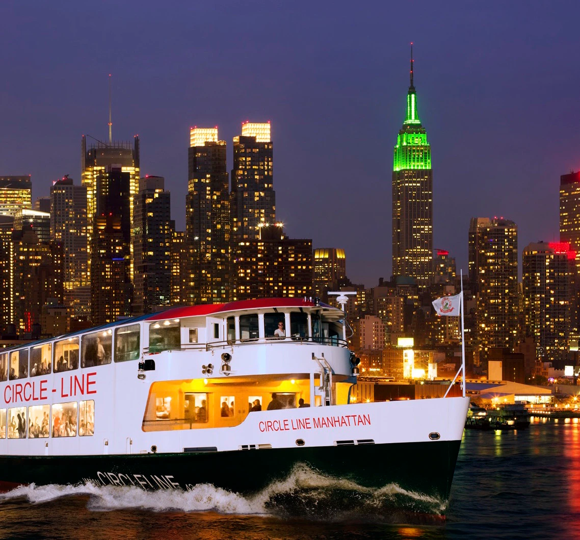 A cruise boat labeled "Circle Line Manhattan" travels on water with the NYC skyline and illuminated Empire State Building in the background.