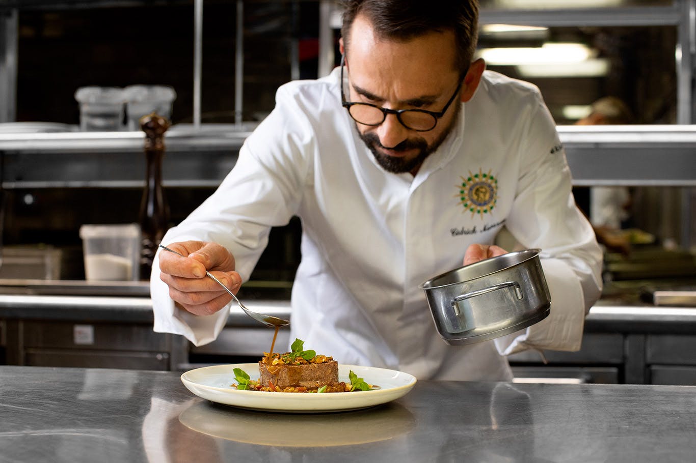 Šéfkuchař v bílé uniformě pečlivě pokovuje talíř v profesionální kuchyni, drží hrnec a lžíci.