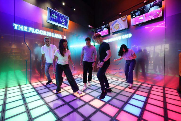 A group of people playing a game on a glowing, colorful floor with "The Floor is Lava" sign in a lively, neon-lit room.