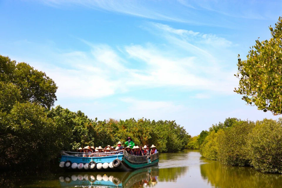 A group of people wearing hats sitting on two boats in a narrow river surrounded by green trees under a blue sky.