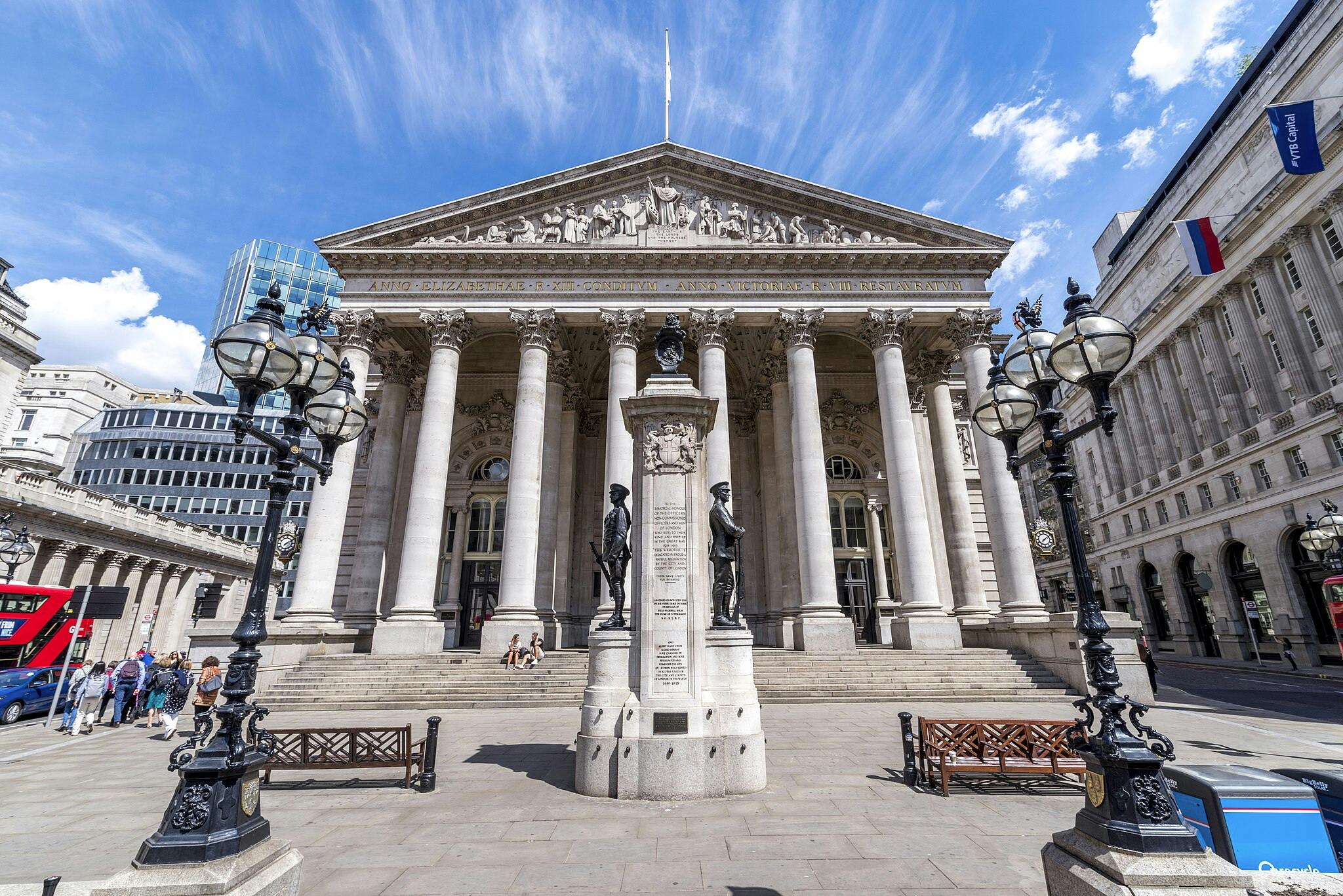 Neoklasyczny budynek z kolumnami i posągami, otoczony ozdobnymi latarniami ulicznymi i ławkami. Ludzie stoją z przodu i na schodach.