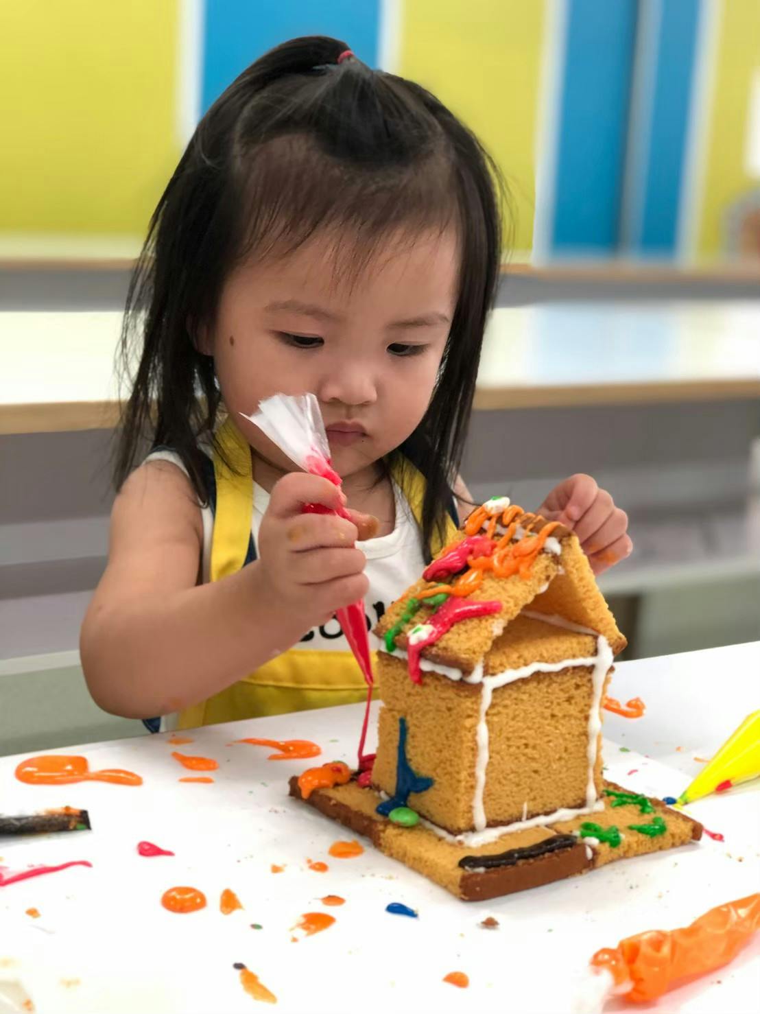 Young child decorating a gingerbread house with colorful icing, in a bright room with shelves in the background.