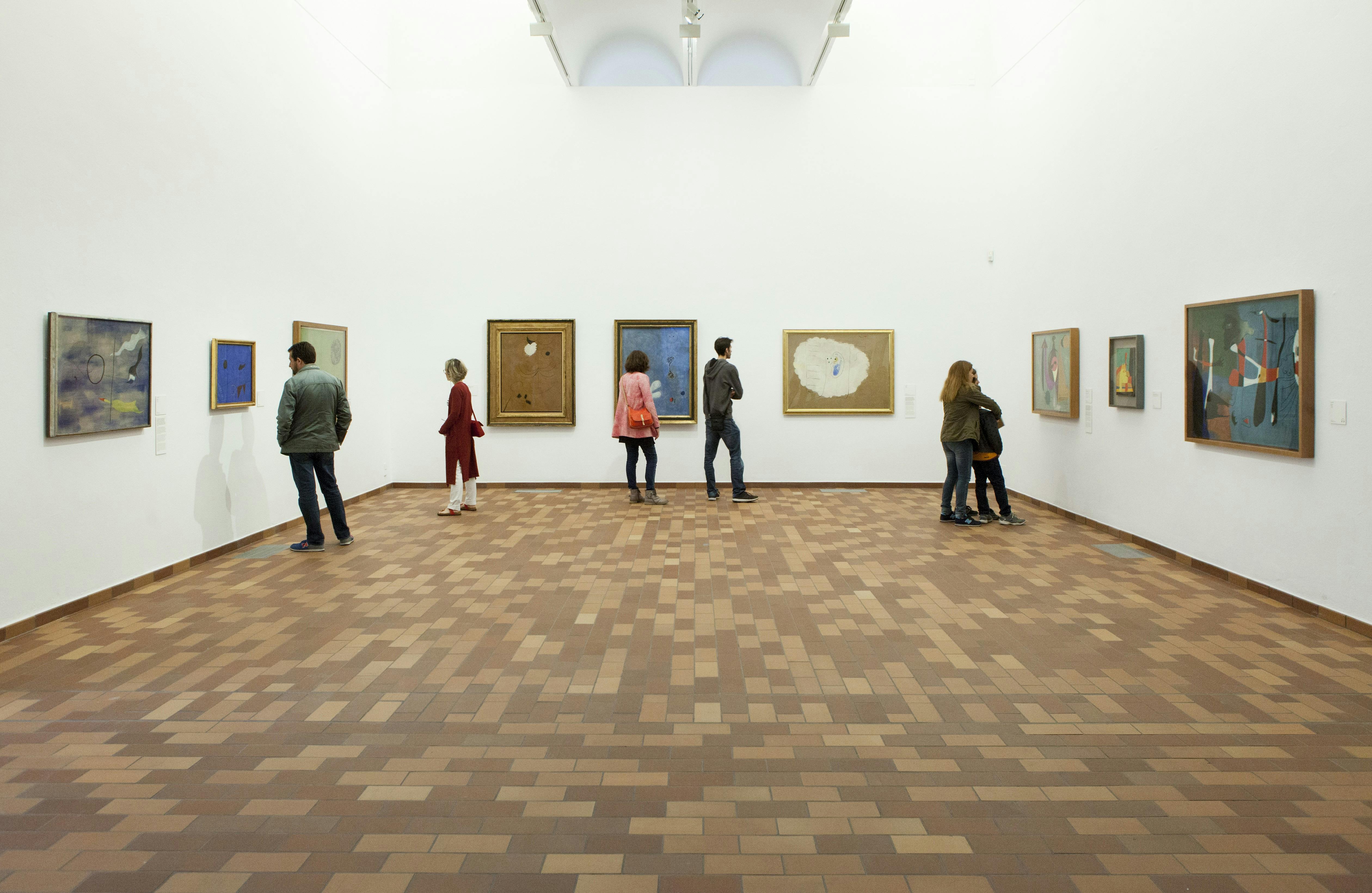 People are observing various paintings in a well-lit art gallery with a brown tiled floor and white walls.