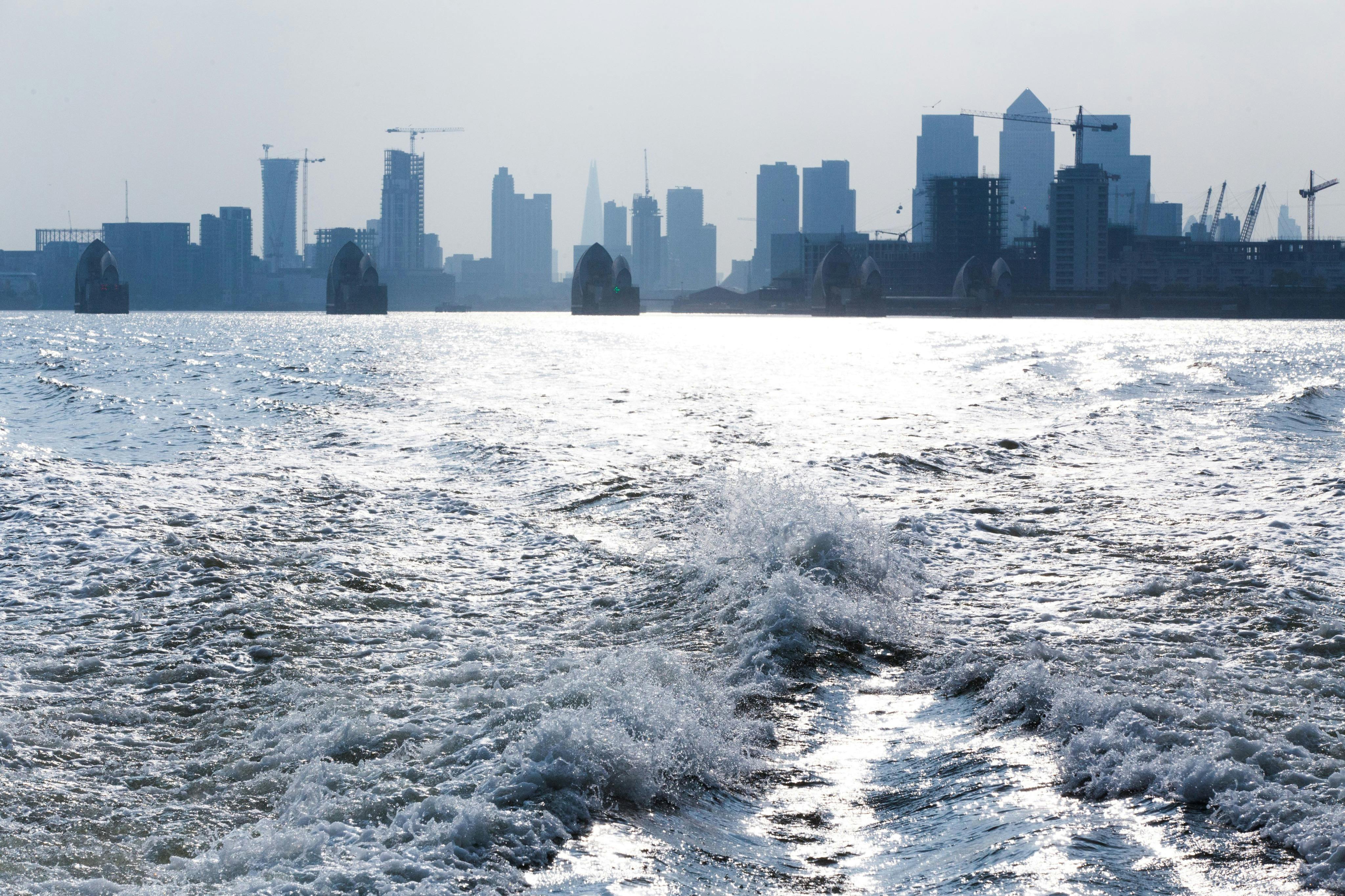 View of a river with waves in the foreground and a cityscape with skyscrapers and cranes in the background.