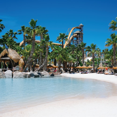 Palme, una piscina simile a una laguna, una spiaggia sabbiosa, capanne di paglia, sedie a sdraio e un alto scivolo d'acqua sotto un cielo azzurro e limpido.