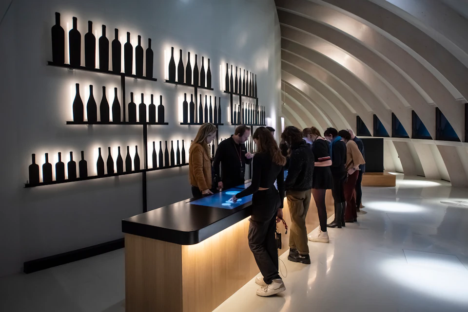 People examining a touch screen at a modern wine tasting room with illuminated bottle silhouettes on the walls.