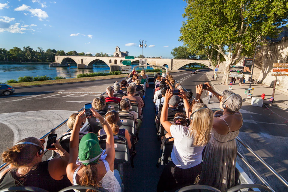Tourists on an open-top bus take photos of a historic bridge over a river on a sunny day.