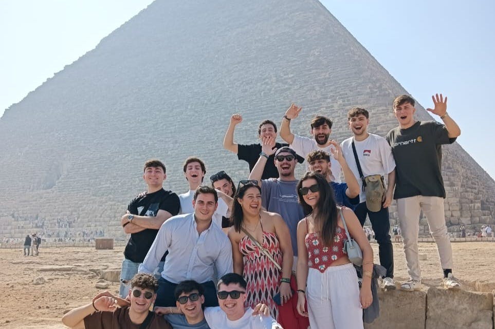 Μια ομάδα ανθρώπων που χαμογελούν και ποζάρουν μπροστά από μια πυραμίδα κάτω από έναν καταγάλανο ουρανό.