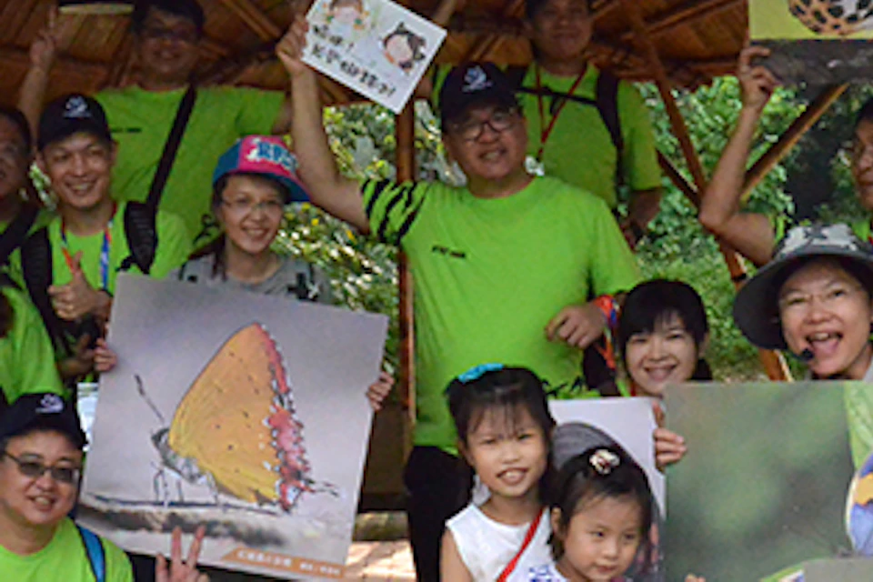 A group of people wearing green shirts holding butterfly posters, posing under a bamboo structure with a woven roof.