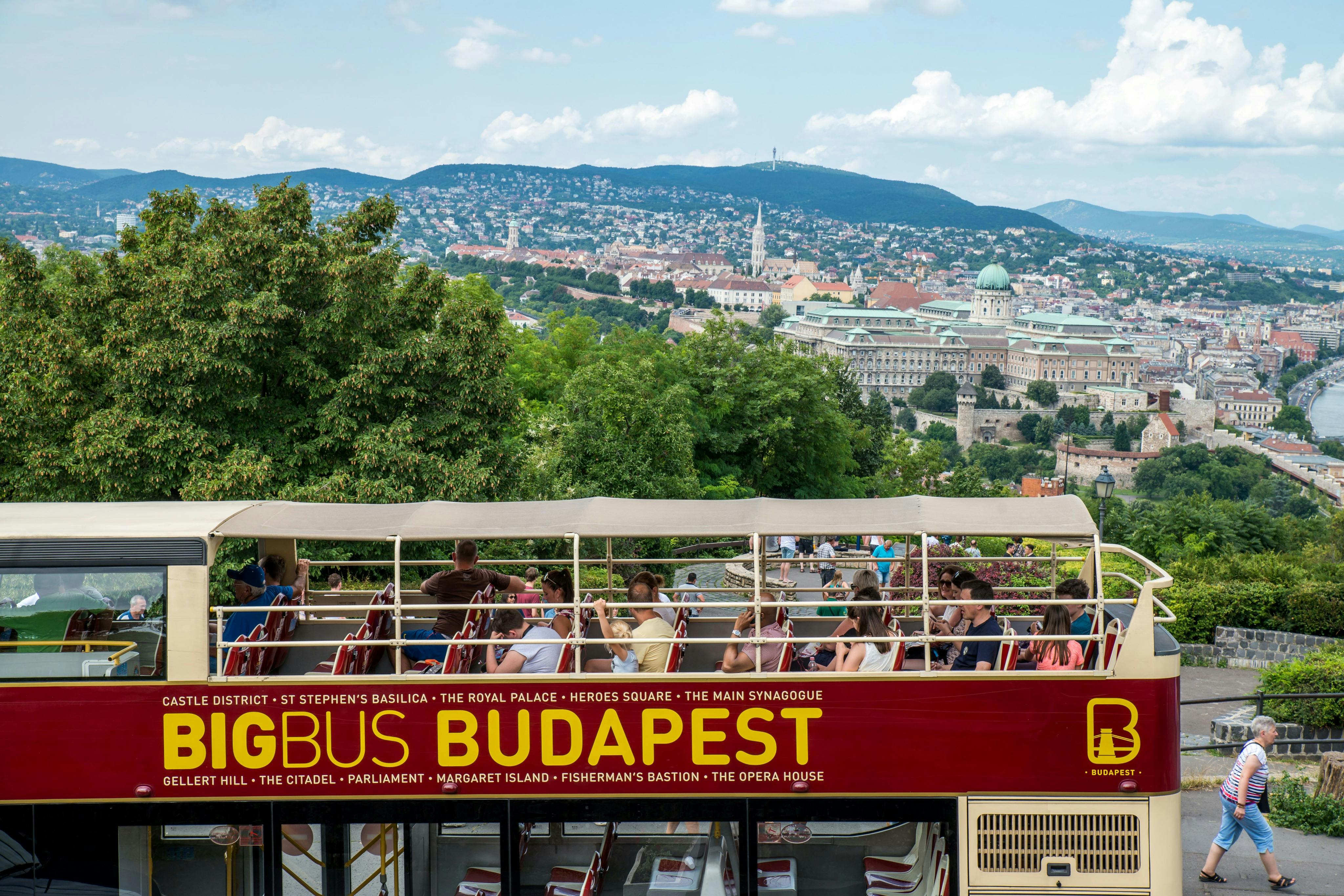 Un bus touristique à deux étages à Budapest avec des touristes assis, surplombant la ligne d'horizon de la ville et les bâtiments historiques.