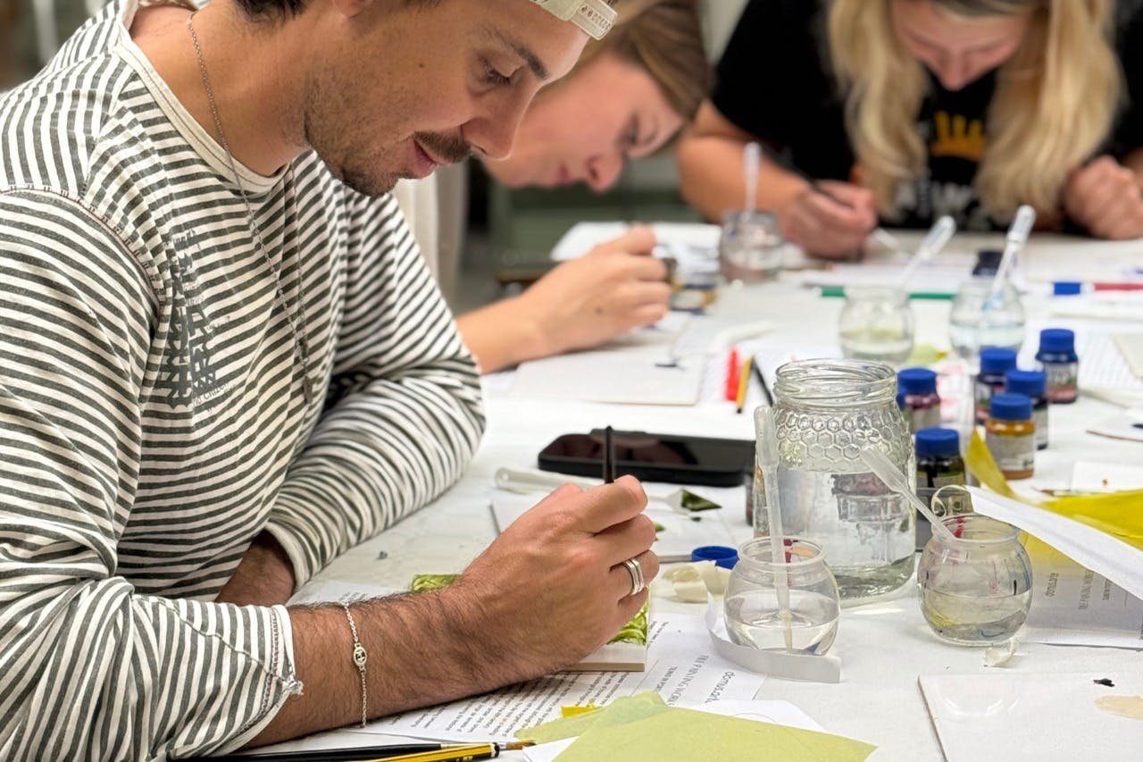 Persone sedute a un tavolo che si concentrano su lavori artistici o di scrittura dettagliati, con vari materiali e strumenti sul tavolo.