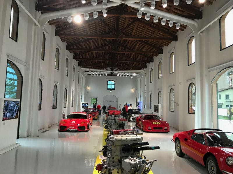 Une salle d'exposition spacieuse aux murs blancs et au plafond en bois présente des voitures de sport rouges et des moteurs de voiture. Plusieurs personnes sont présentes.