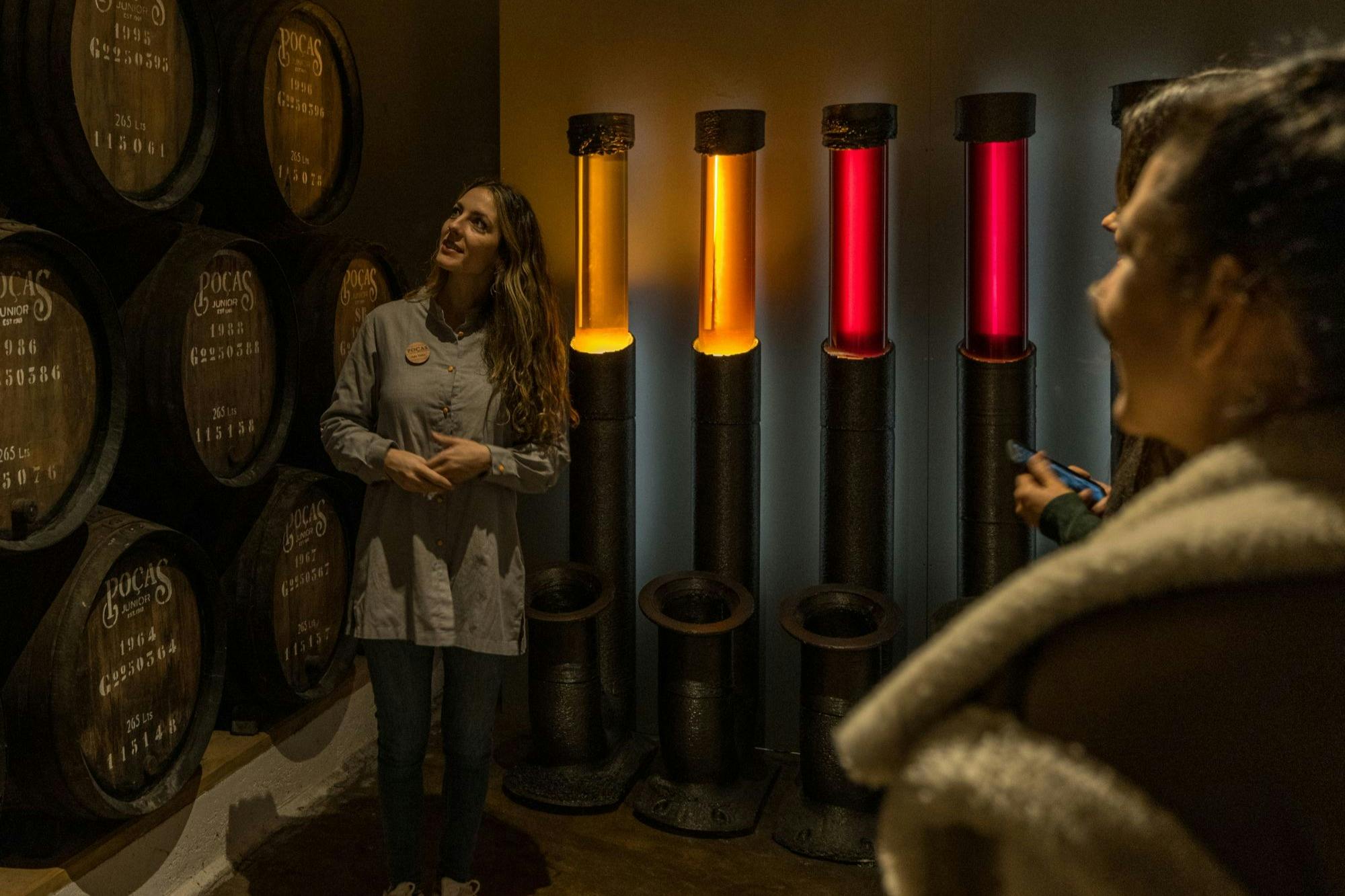 A woman stands near illuminated vertical tubes in red and orange, with barrels in the background in a dimly lit room.