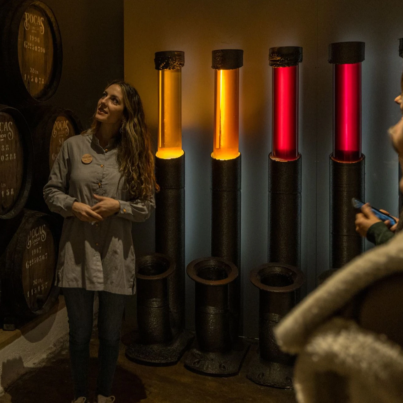 A woman stands near illuminated vertical tubes in red and orange, with barrels in the background in a dimly lit room.