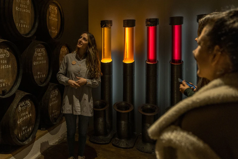 A woman stands near illuminated vertical tubes in red and orange, with barrels in the background in a dimly lit room.