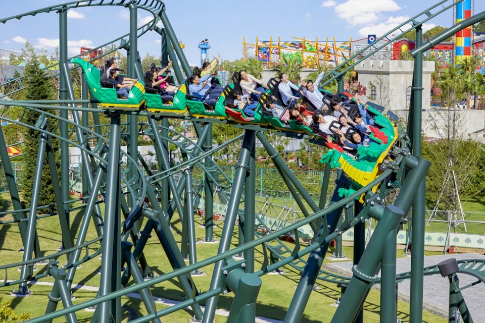 People riding a colorful rollercoaster with green supports in an amusement park on a sunny day.