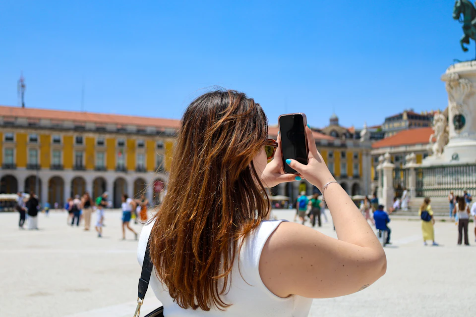 Person with long hair, wearing a white tank top, taking a photo with a smartphone in a sunny town square with historic buildings.