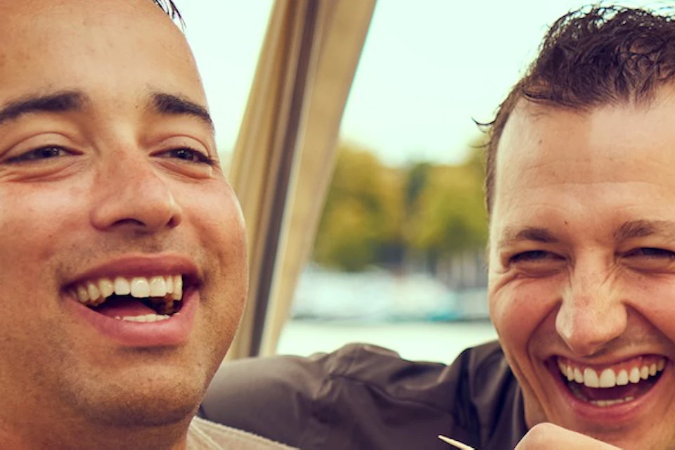 Two men sitting on a boat, laughing, one holding a glass of wine, with a blurred water and tree background.