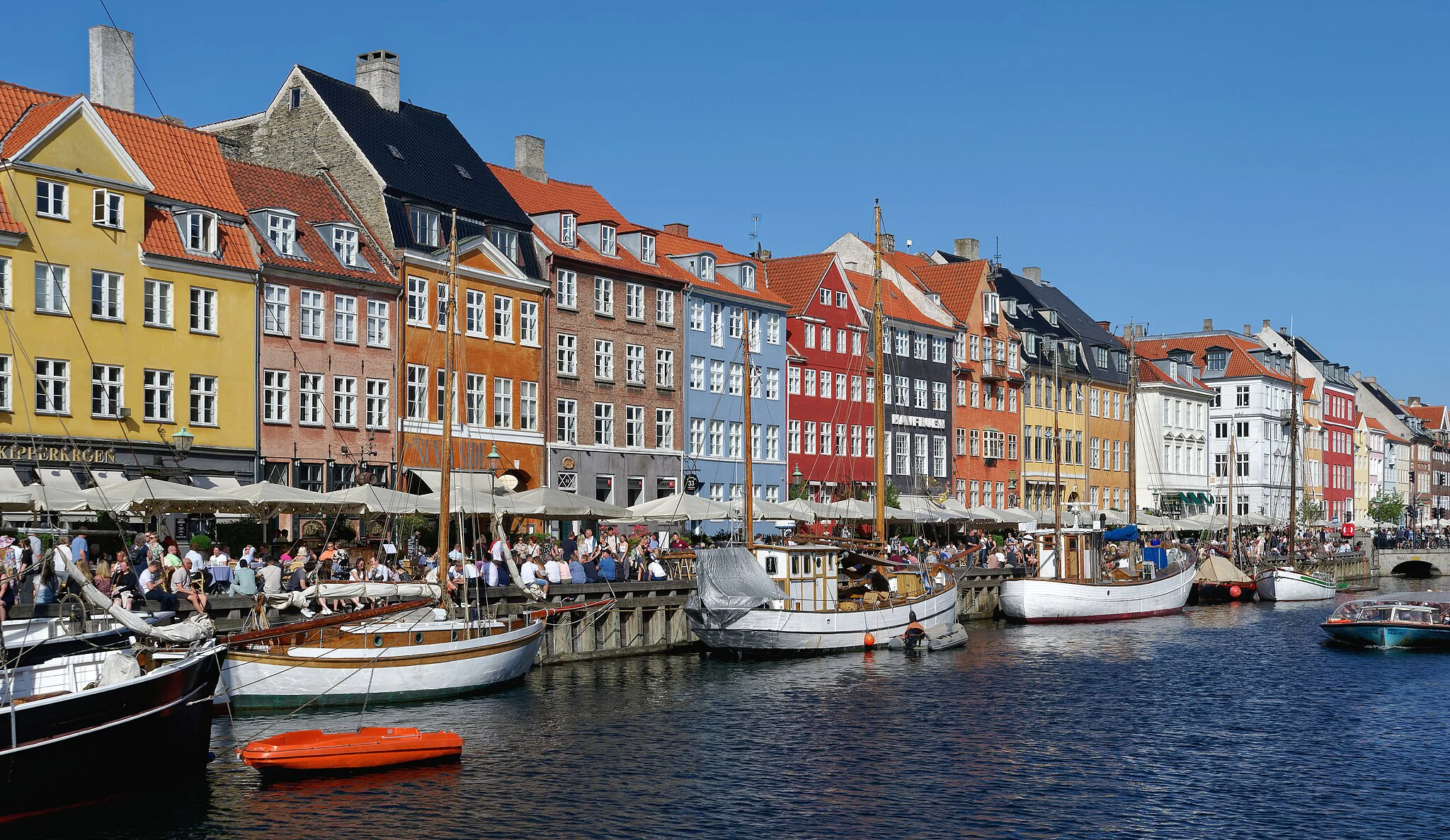 Des bâtiments colorés bordent un front de mer bondé de gens, de bateaux et de cafés en plein air sous un ciel bleu limpide.