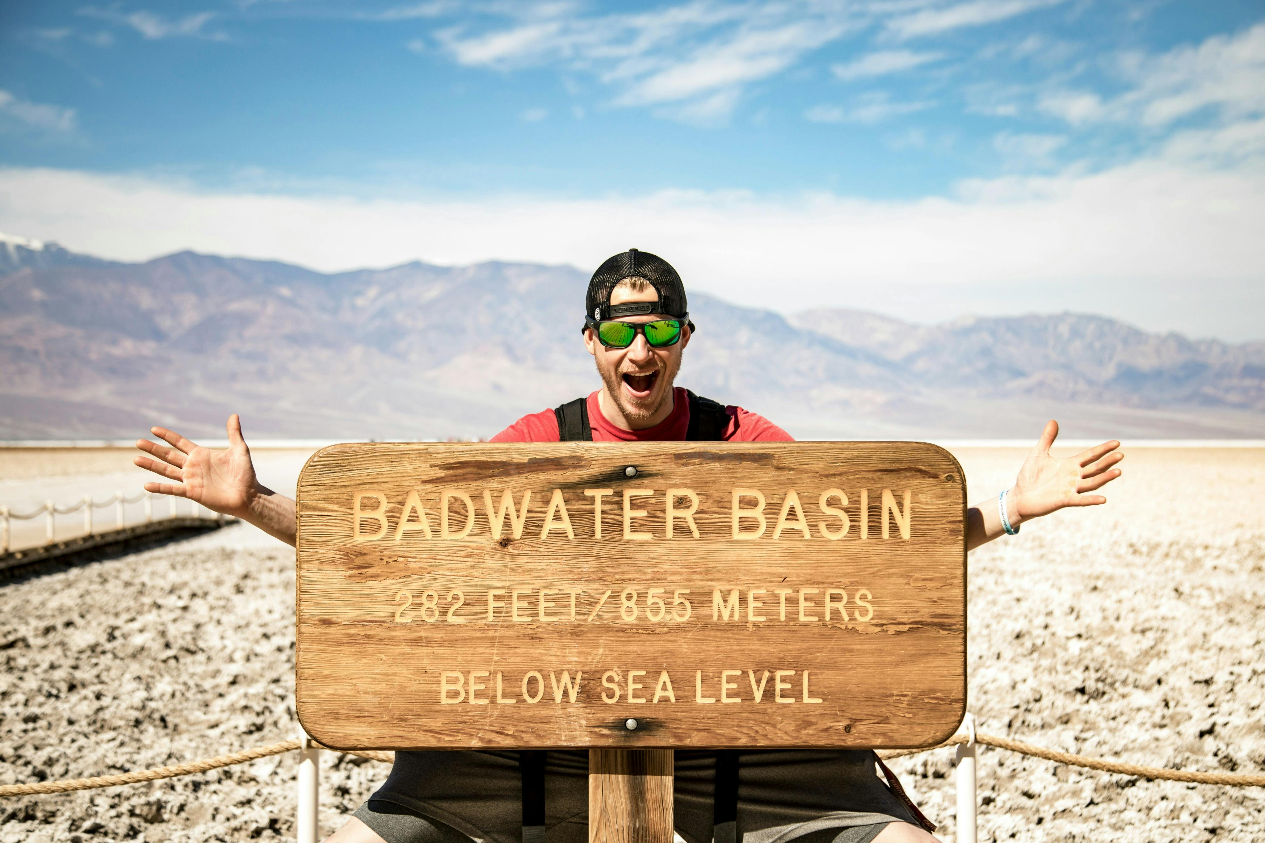A person is posing behind the Badwater Basin sign in a desert landscape, with mountains and a blue sky in the background.