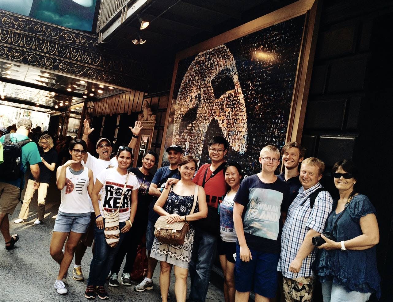A group of people pose and smile in front of a theater entrance with a mosaic artwork of a mask in the background.