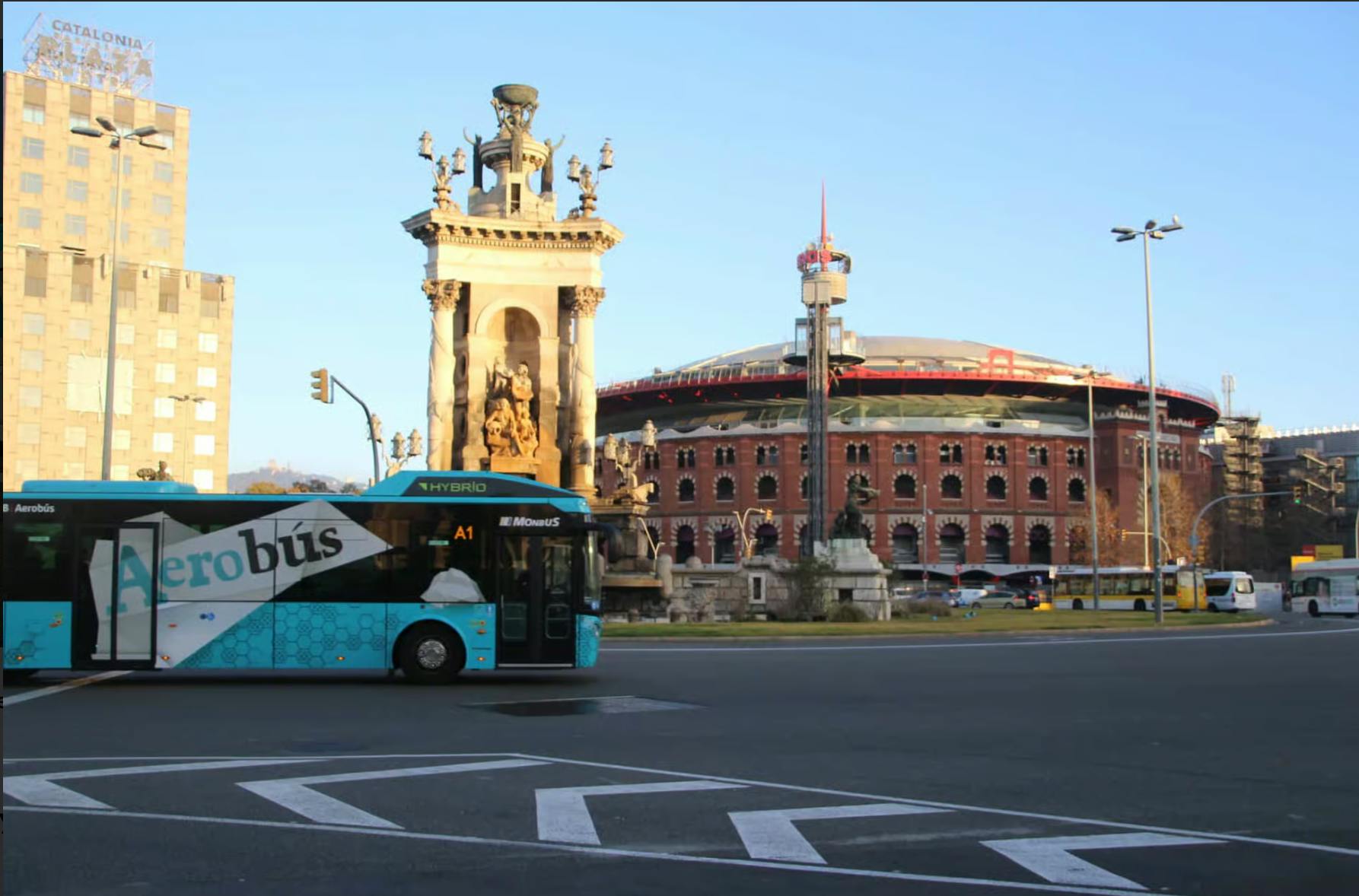 Un bus hybride passe devant un monument historique et un grand bâtiment circulaire en briques par une journée ensoleillée.
