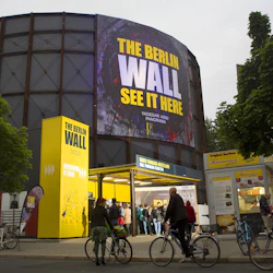 Människor på cyklar och fotgängare står utanför en byggnad med en stor display som annonserar "Berlinmuren" panoramainstallation.