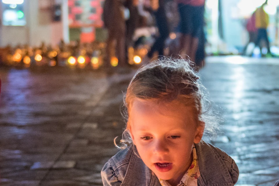 Children crouch to admire lit lanterns on a cobblestone street at night, with a busy, illuminated background.