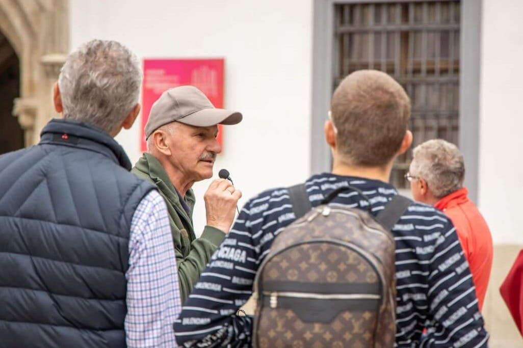 Ένας ηλικιωμένος άνδρας με καπέλο κρατά ένα μικρόφωνο, απευθυνόμενος σε δύο άνδρες με casual ρούχα, ο ένας με σακίδιο, σε υπαίθριο χώρο.