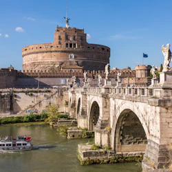 En båt på en flod passerar under en gammal stenbro prydd med statyer, med en fästningsliknande cirkulär byggnad i bakgrunden.