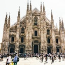 Folla di persone che cammina davanti a una grande cattedrale gotica con guglie intricate e architettura dettagliata.
