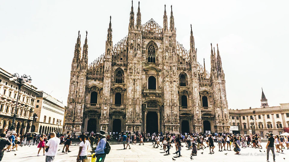 Crowd in front of an ornate Gothic cathedral with intricate spires and detailed facade on a sunny day.