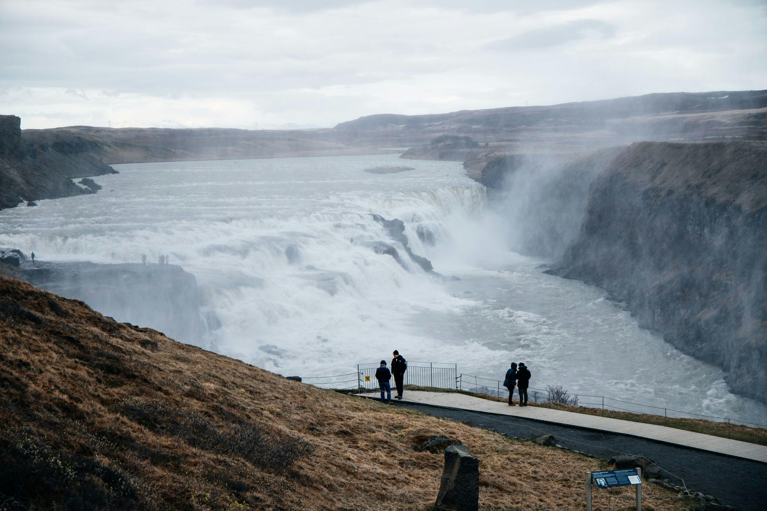People standing on a viewing platform overlooking a large waterfall surrounded by rugged terrain under an overcast sky.