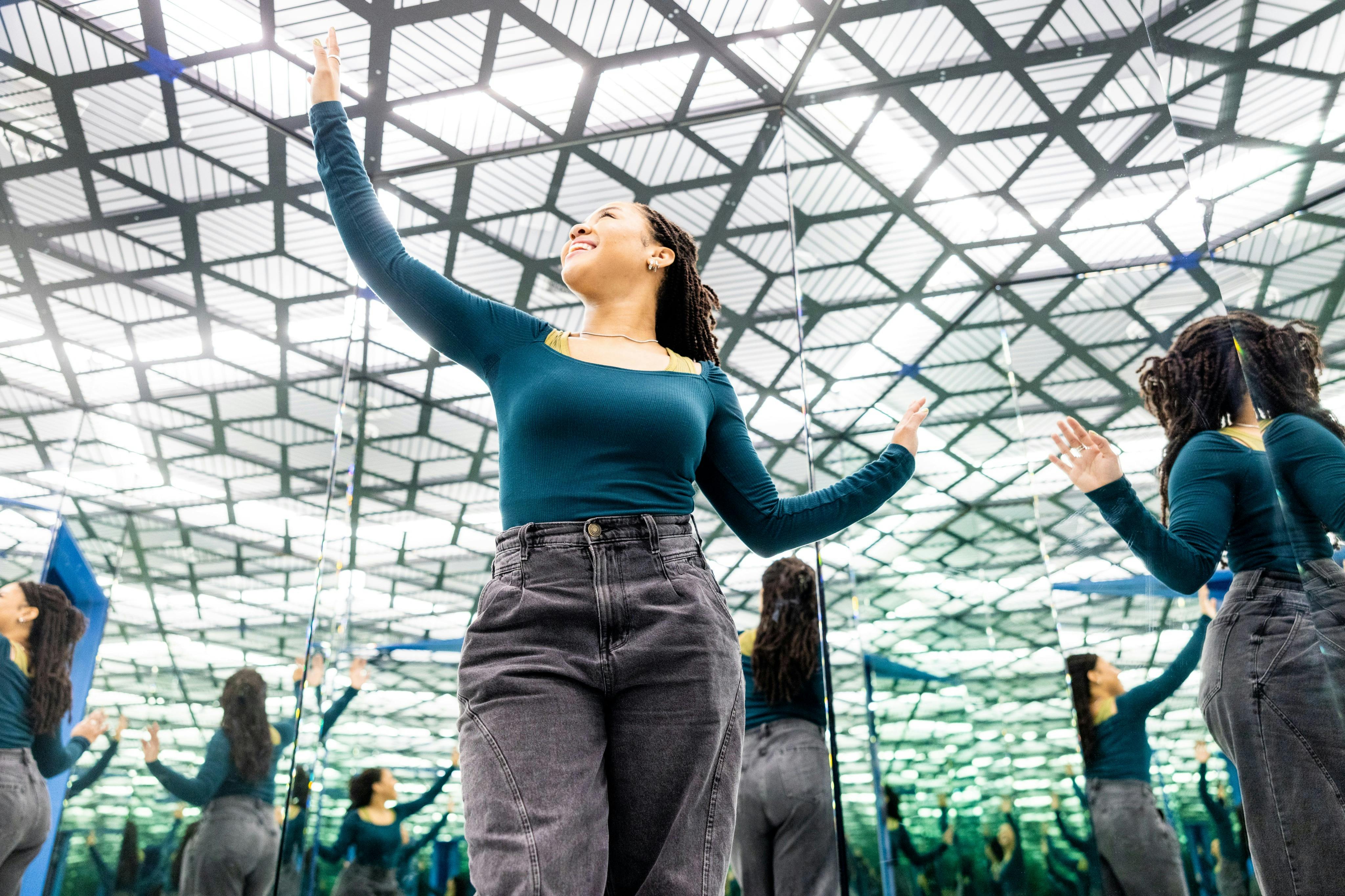 Μια γυναίκα με μπλε μπλούζα και γκρι παντελόνι στέκεται σε ένα δωμάτιο με καθρέφτες, δημιουργώντας πολλαπλές αντανακλάσεις.