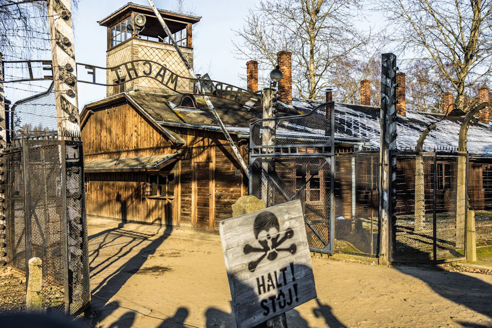 A historical site with a wooden building, barbed wire fencing, and a sign reading "Halt! Stoj!" featuring a skull and crossbones.
