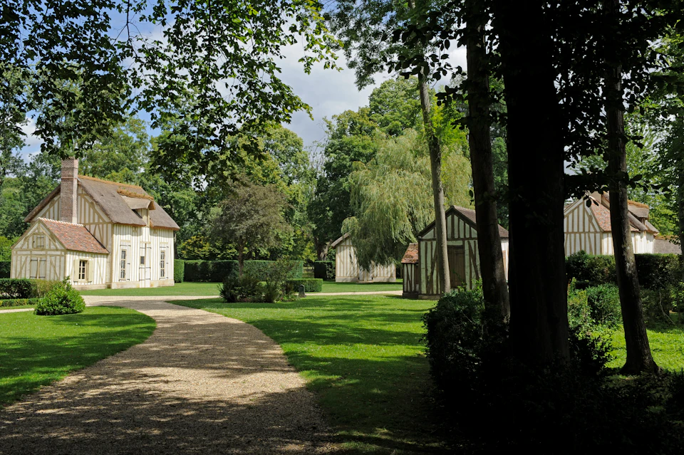 A shaded gravel path curves through a grassy area with several wooden cottages surrounded by trees under a partly cloudy sky.