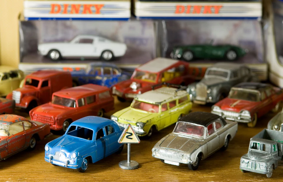Assorted vintage toy cars in various colors on a wooden surface with a small road sign and boxed toy cars in the background.