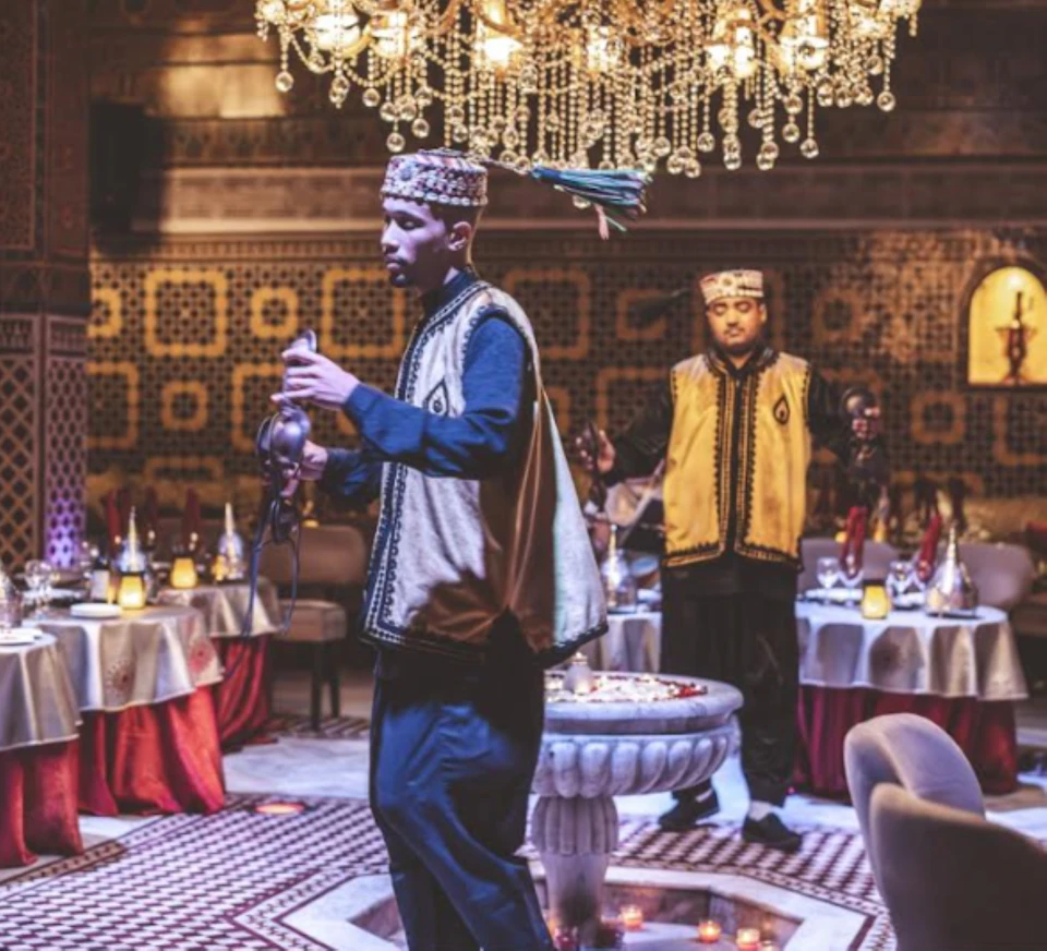 Two men wearing traditional attire serve tea in an ornate restaurant with chandeliers and patterned walls.