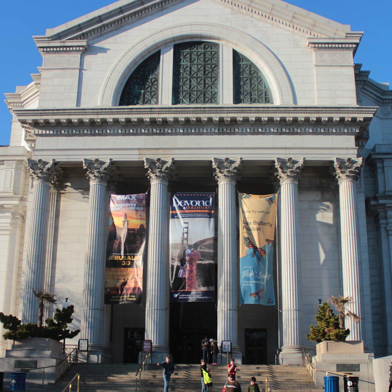 A large neoclassical building with columns, banners, and a large arched window, with people in front on the steps.