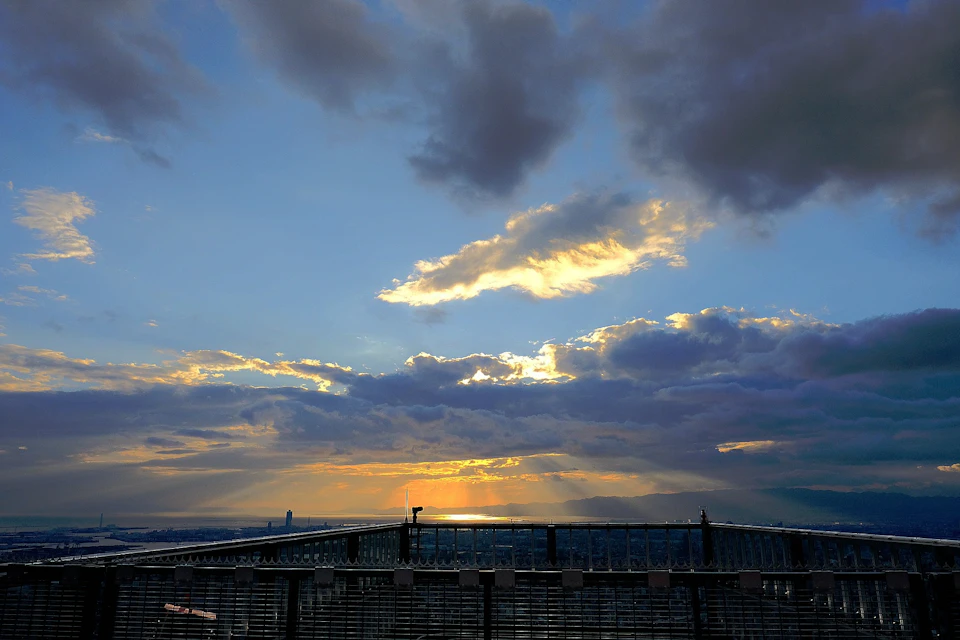 Sunset view from a rooftop with dramatic clouds and sun rays piercing through, overlooking a distant city skyline.