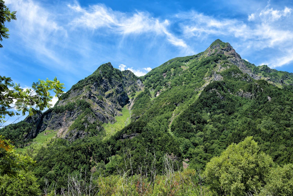 A lush green mountain under a bright blue sky with scattered white clouds, surrounded by forested terrain.