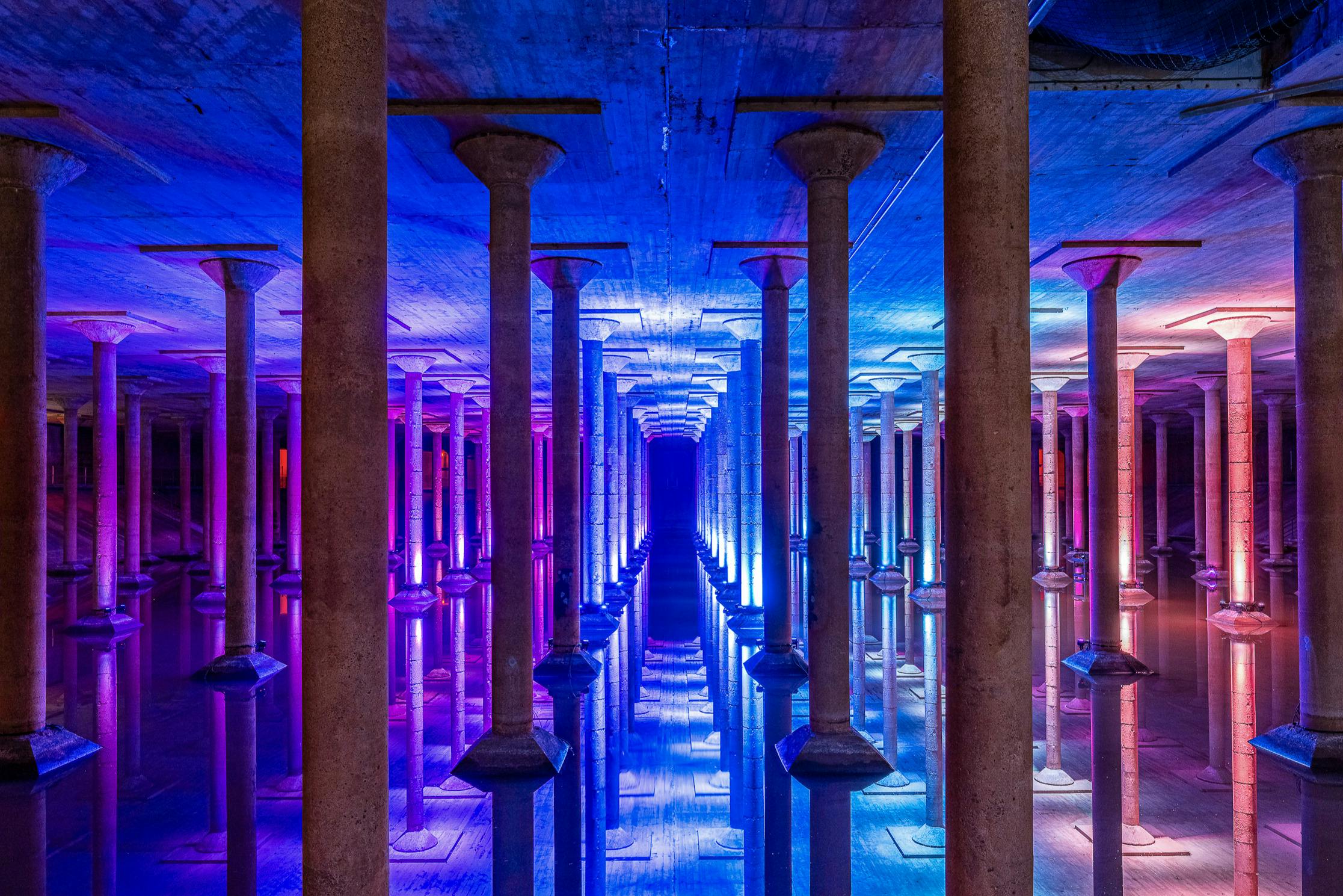 선명한 파란색과 보라색 불빛이 비추는 석조 기둥이 있는 거울로 장식된 복도는 대칭적이고 반사되는 장면을 연출합니다.