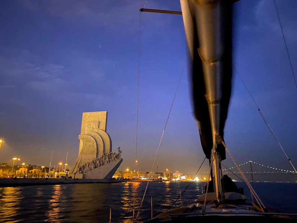 A sailboat on water at dusk with a large monument of figures on a curved structure in the background, illuminated by city lights.