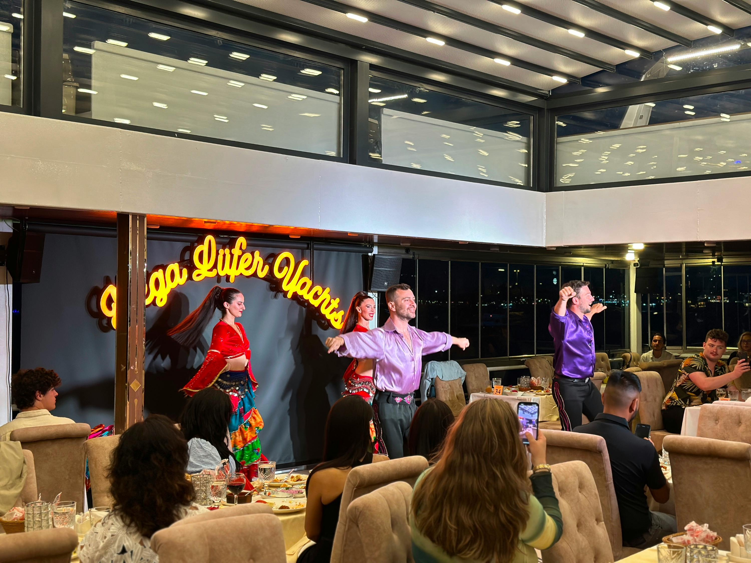 Performers in colorful costumes dance on a stage in front of an audience in a well-lit indoor venue.