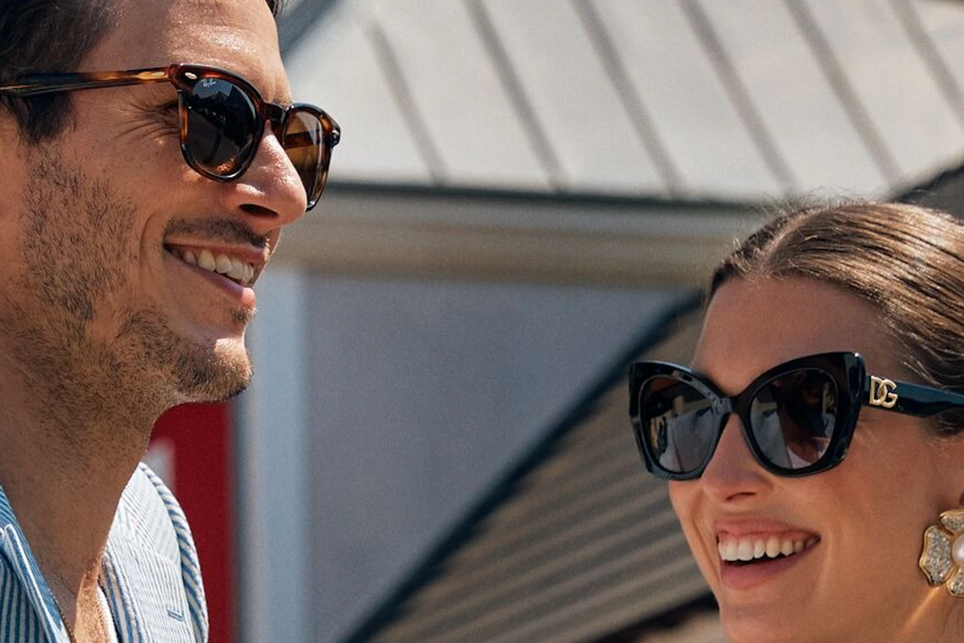 A man and woman in stylish outfits laugh outside La Vallée Village shopping center on a sunny day.