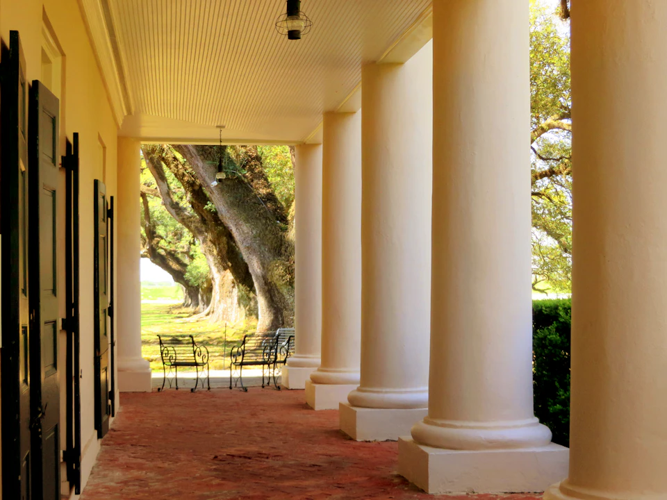 White columns on a porch with wrought-iron chairs, a brick floor, and large trees in the background.