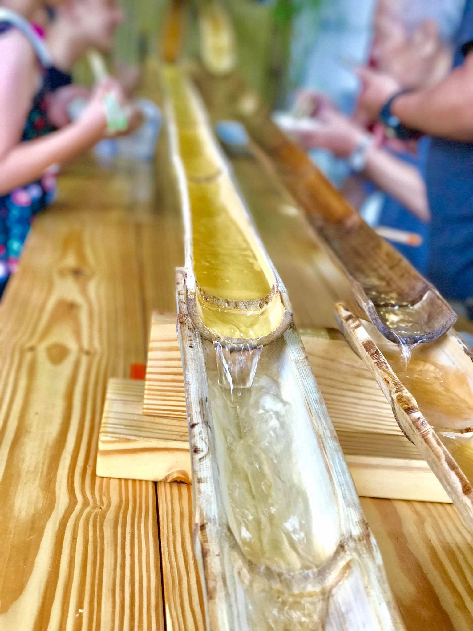 A flowing bamboo water chute on a wooden table, with people in the background holding bowls, likely engaging with the setup.