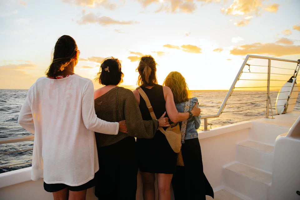 Four people stand on a boat deck at sunset, arms around each other, looking out at the ocean and the colorful sky.